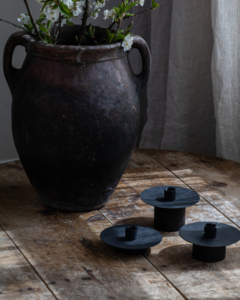 3 stk lysestaker i jern i ulik høyde står på et bord sammen med en stor vintage krukke 