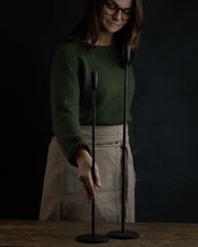 2stk lysestaker i smijern i ulik høyde. En dame i grønn genser holder på den ene lysestaken 